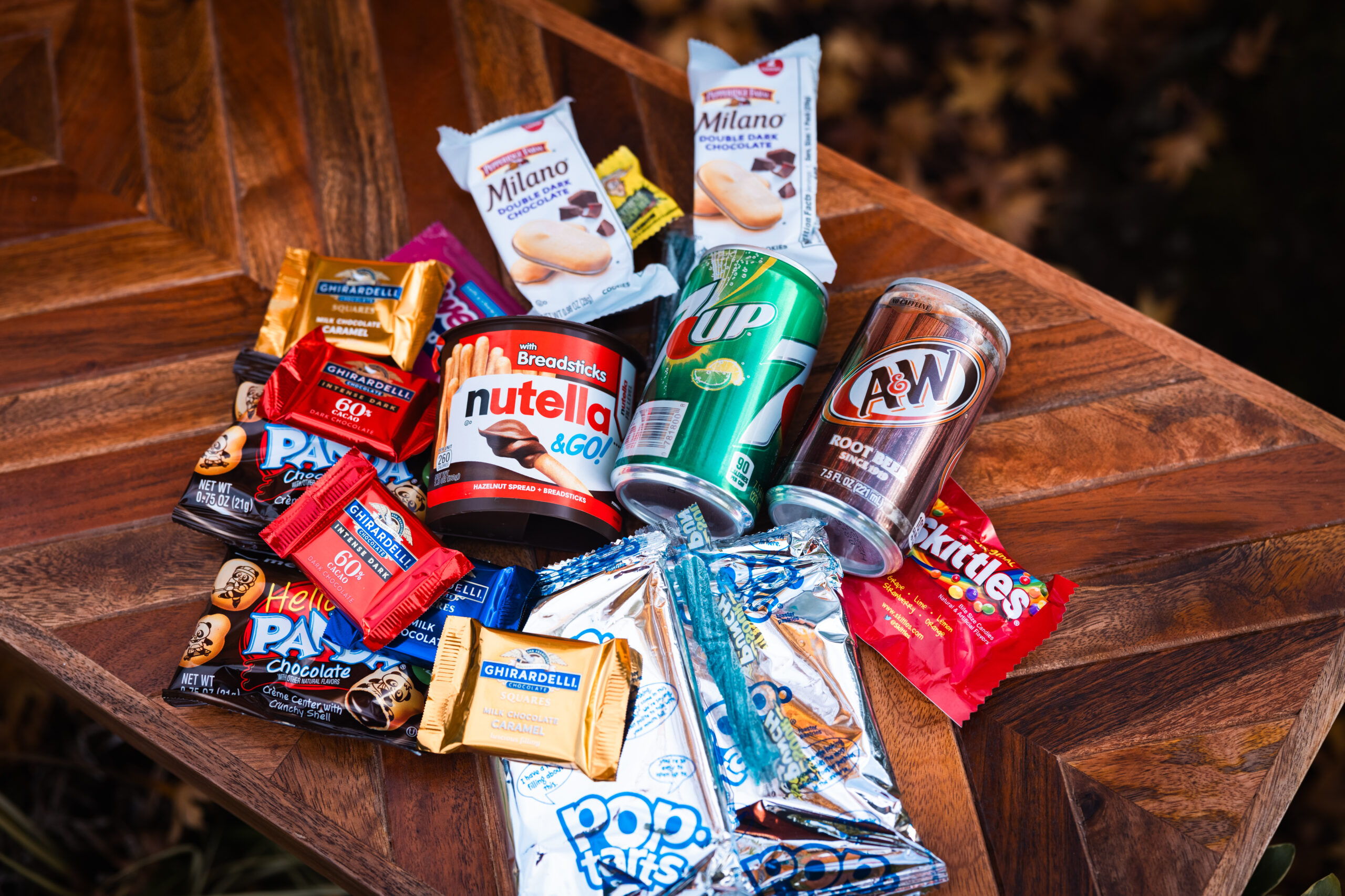 A variety of snacks and drinks on a wooden surface, including Nutella, Milano cookies, Pop-Tarts, Skittles, a 7UP can, an A&W Root Beer can, Hi-Chew candies, Ghirardelli chocolates, and Hello Panda biscuits.