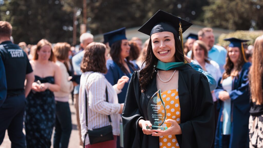A woman in a graduation gown proudly holds an Distinguished Graduate award, celebrating her academic achievement.