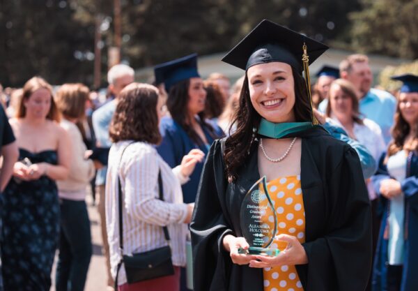 A woman in a graduation gown proudly holds an Distinguished Graduate award, celebrating her academic achievement.