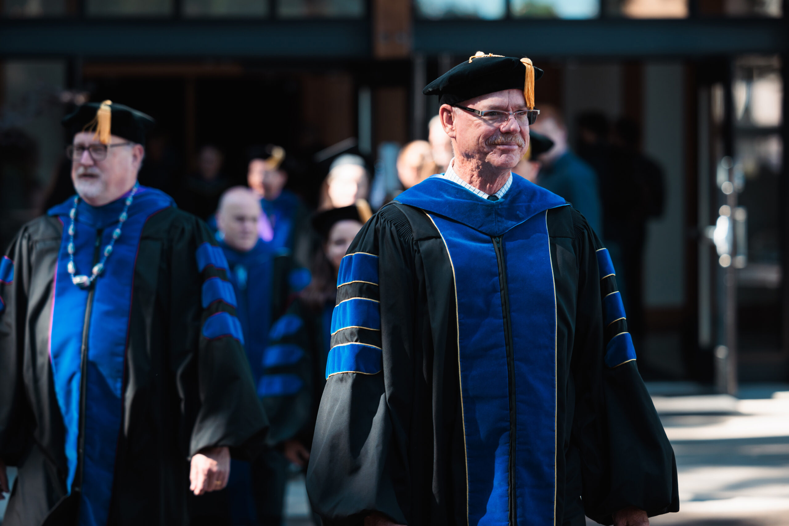 A faculty member in a doctoral-level graduation cap and gown smiles as he exits a building with other faculty following behind.