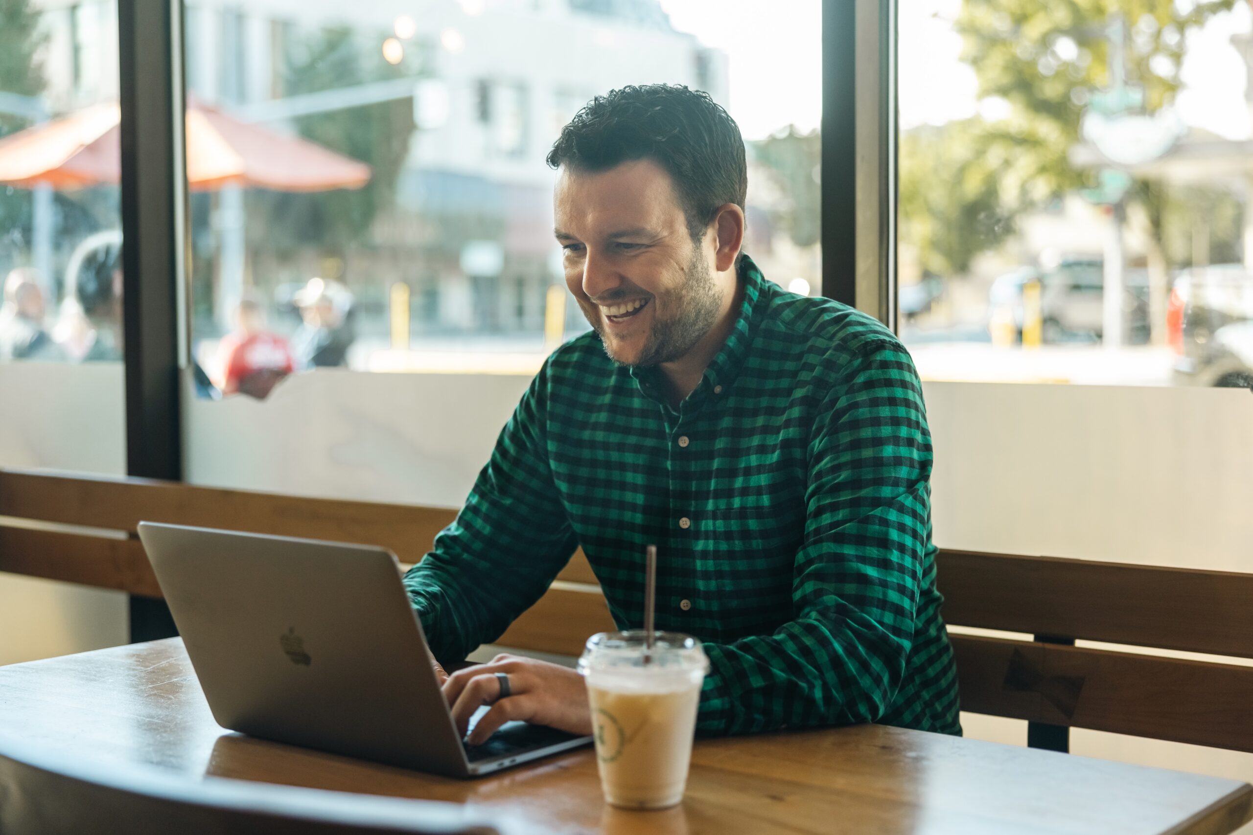 A man in a green checkered shirt is sitting at a wooden table in a café, smiling at his laptop. A cup of iced coffee is on the table. Sunlight filters through large windows behind him, with a street scene outside.