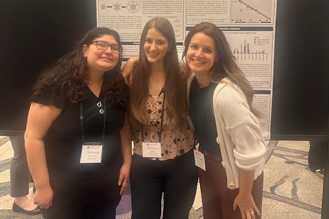 Three people stand together, smiling, in front of a scientific poster. They are wearing name tags and appear to be at a conference or presentation.