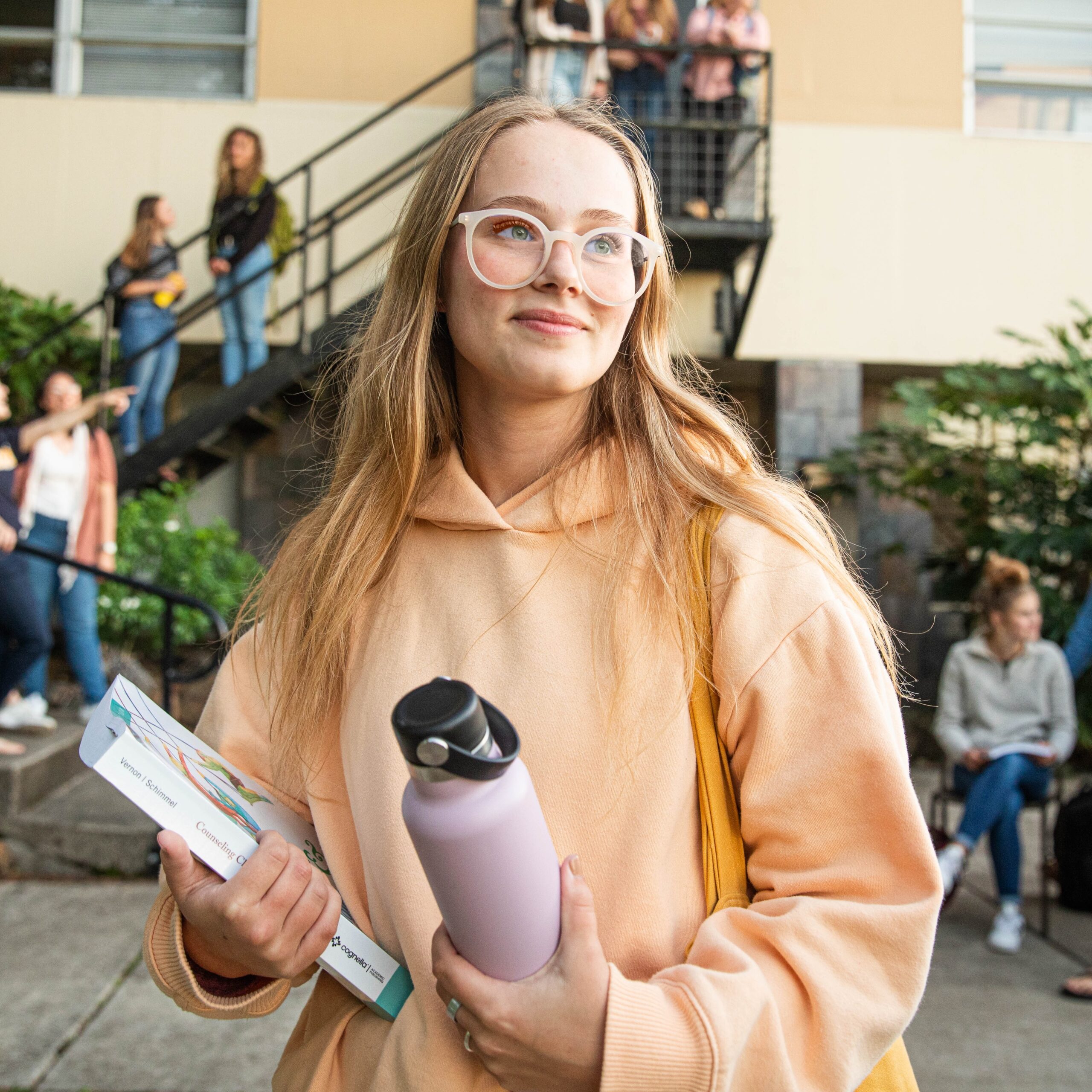 A young woman in glasses stands holding a book and a water bottle, appearing focused and content.