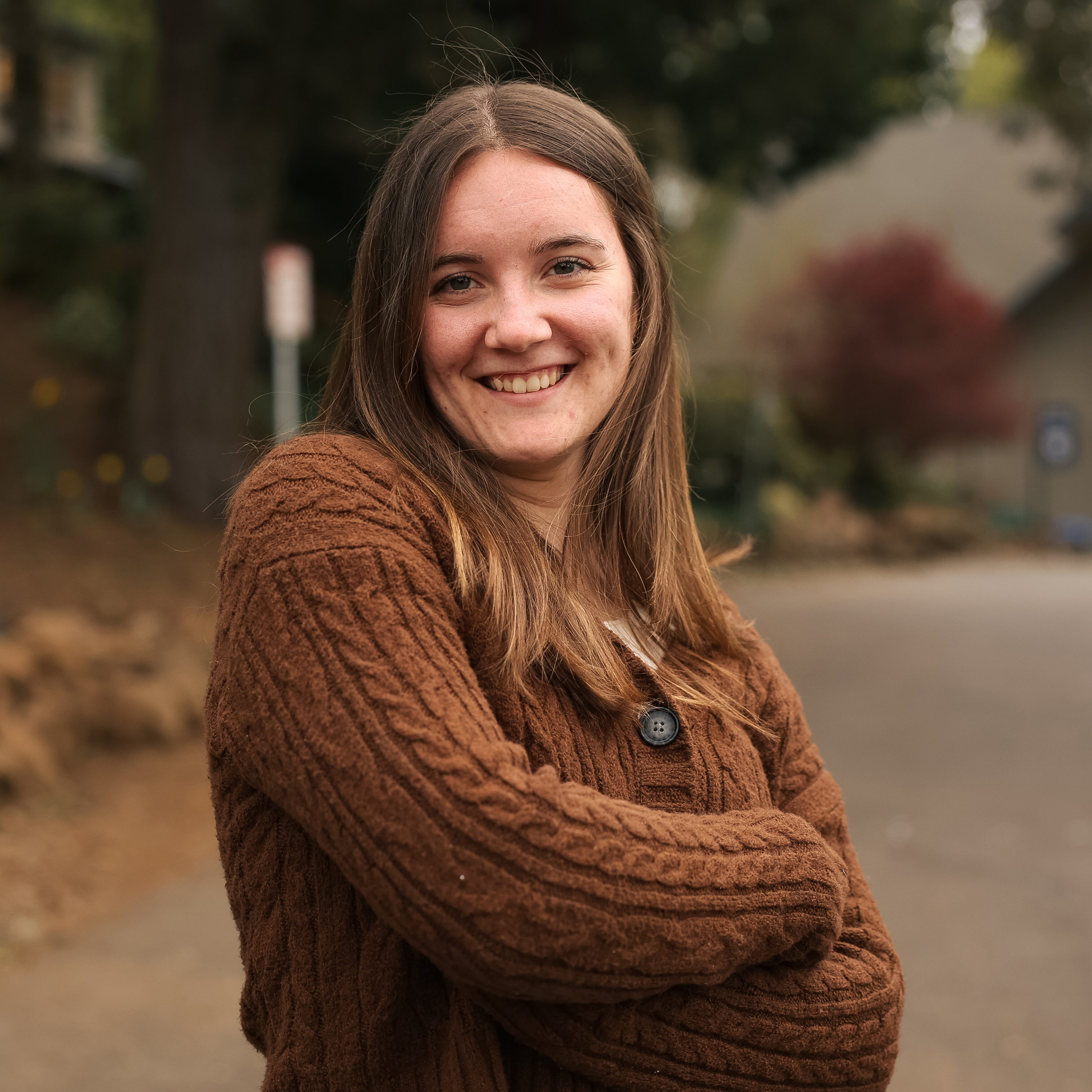 A young woman poses for the camera with her arms crossed and a big smile in an outdoor setting.