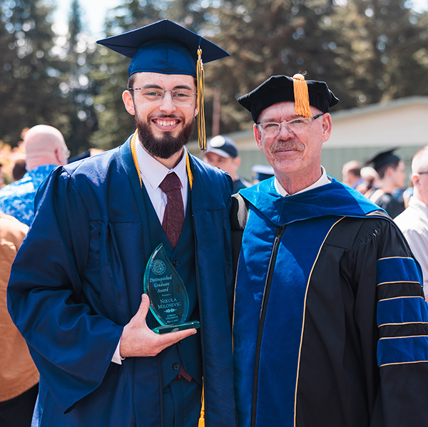 A student and professor stand together and smile in their regalia at a college graduation