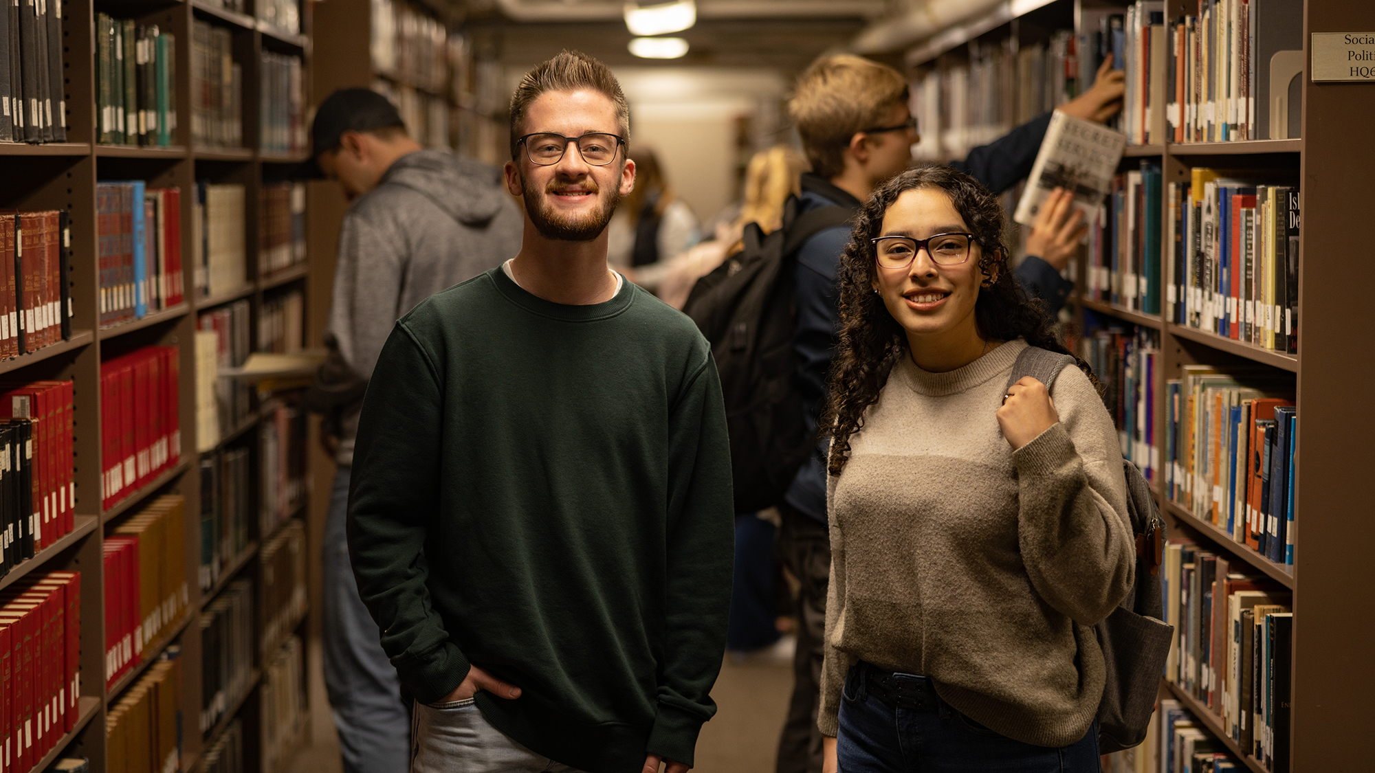 A young man and woman stand in the library and smile at the camera with rows of bookshelves behind them.