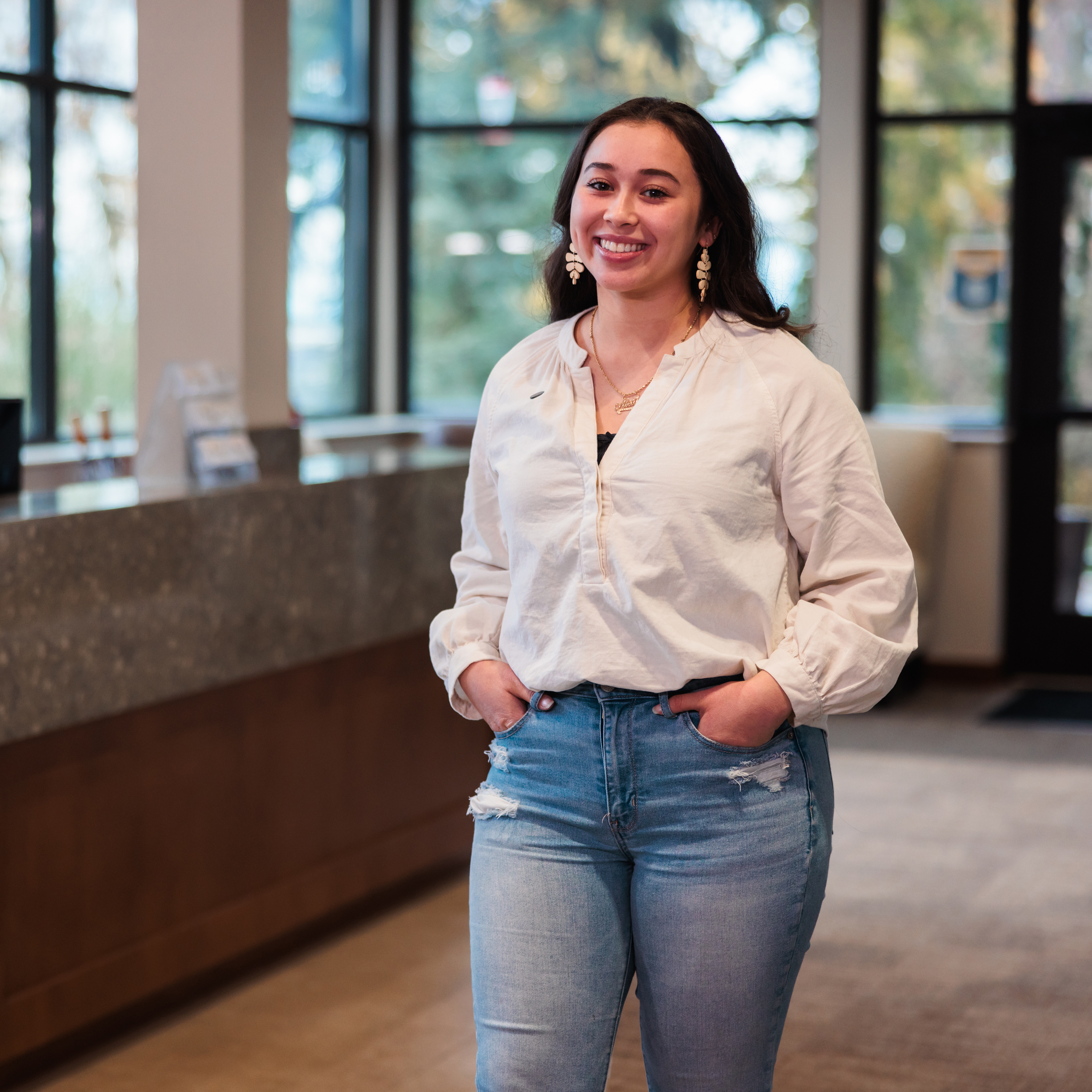A young woman dressed in jeans and a white shirt stands in a bright, welcoming lobby space.