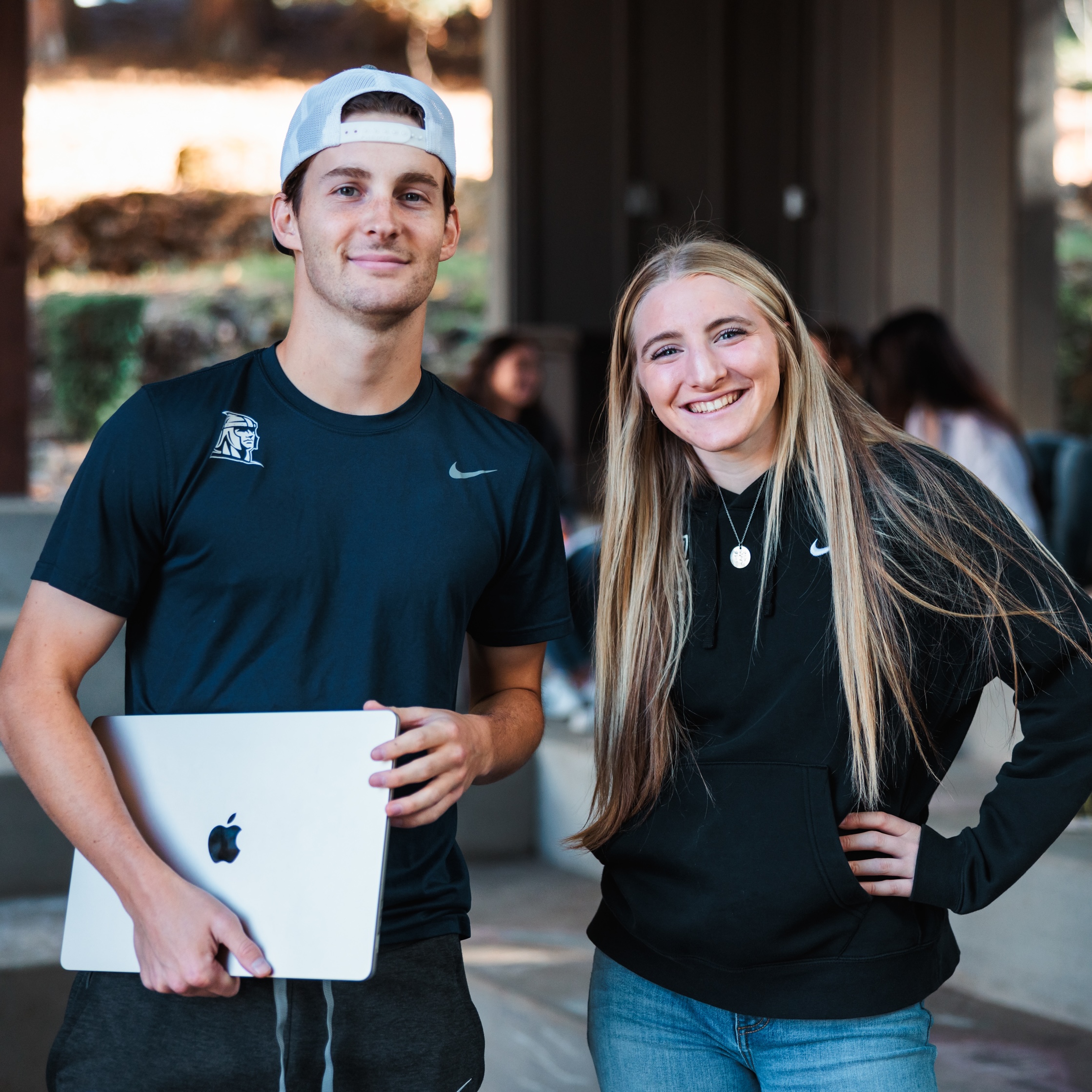 Two students, one holding a laptop, standing side by side, both smiling and dressed casually outdoors.