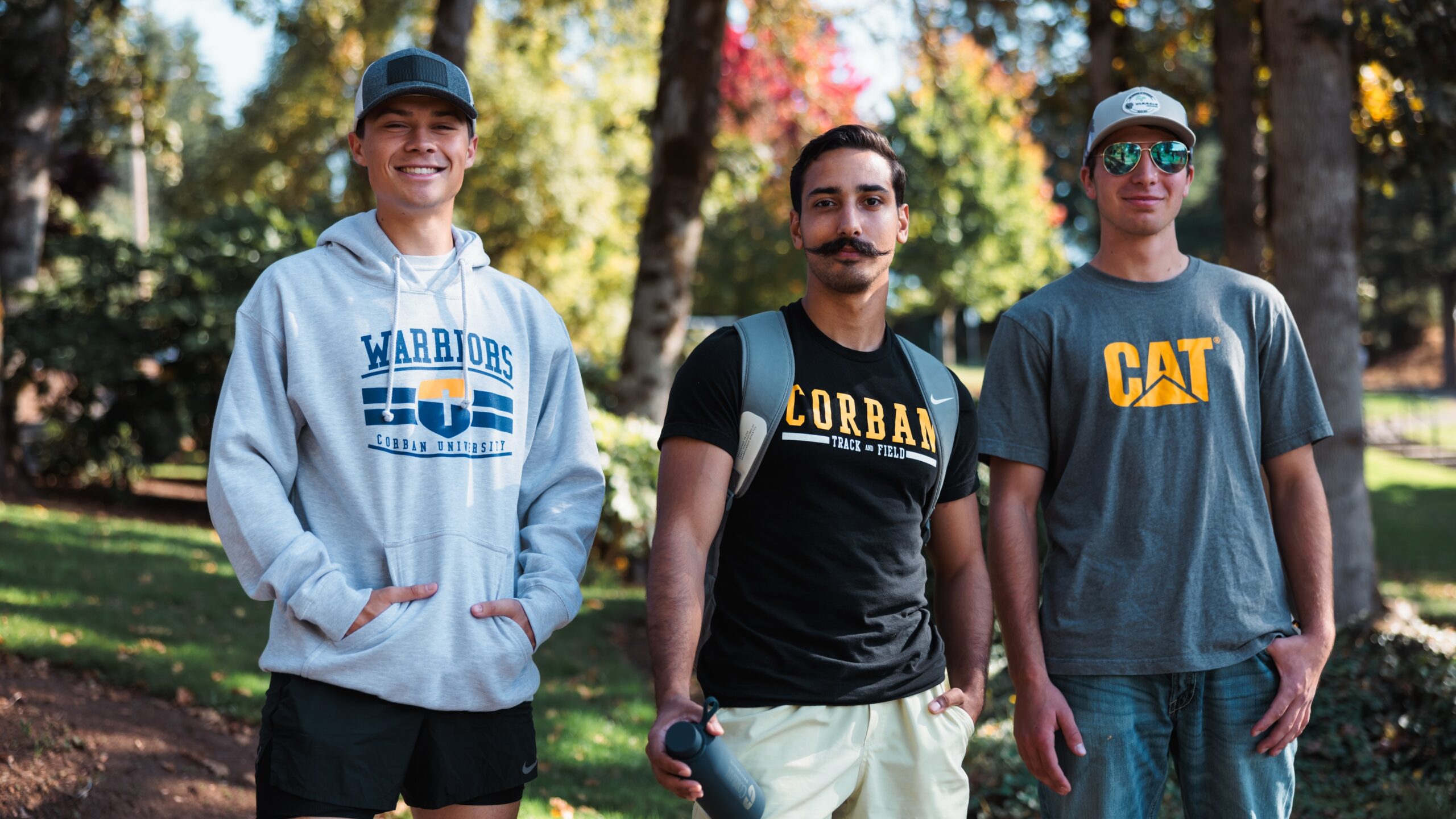 Three young men dressed in casual t-shirts and hoodies, standing together and smiling at the camera.