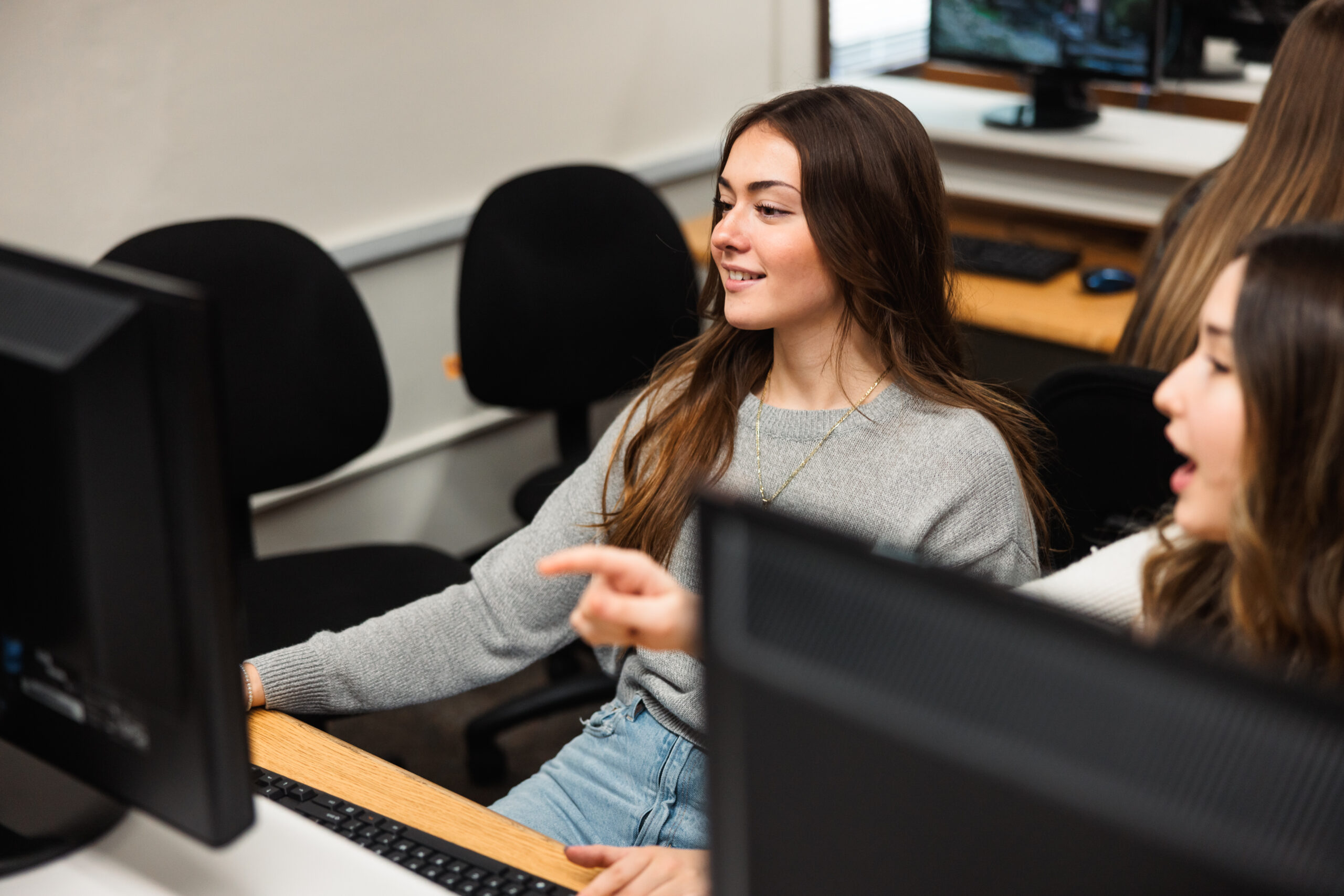Two women engaged in discussion while working at a computer in a computer lab.