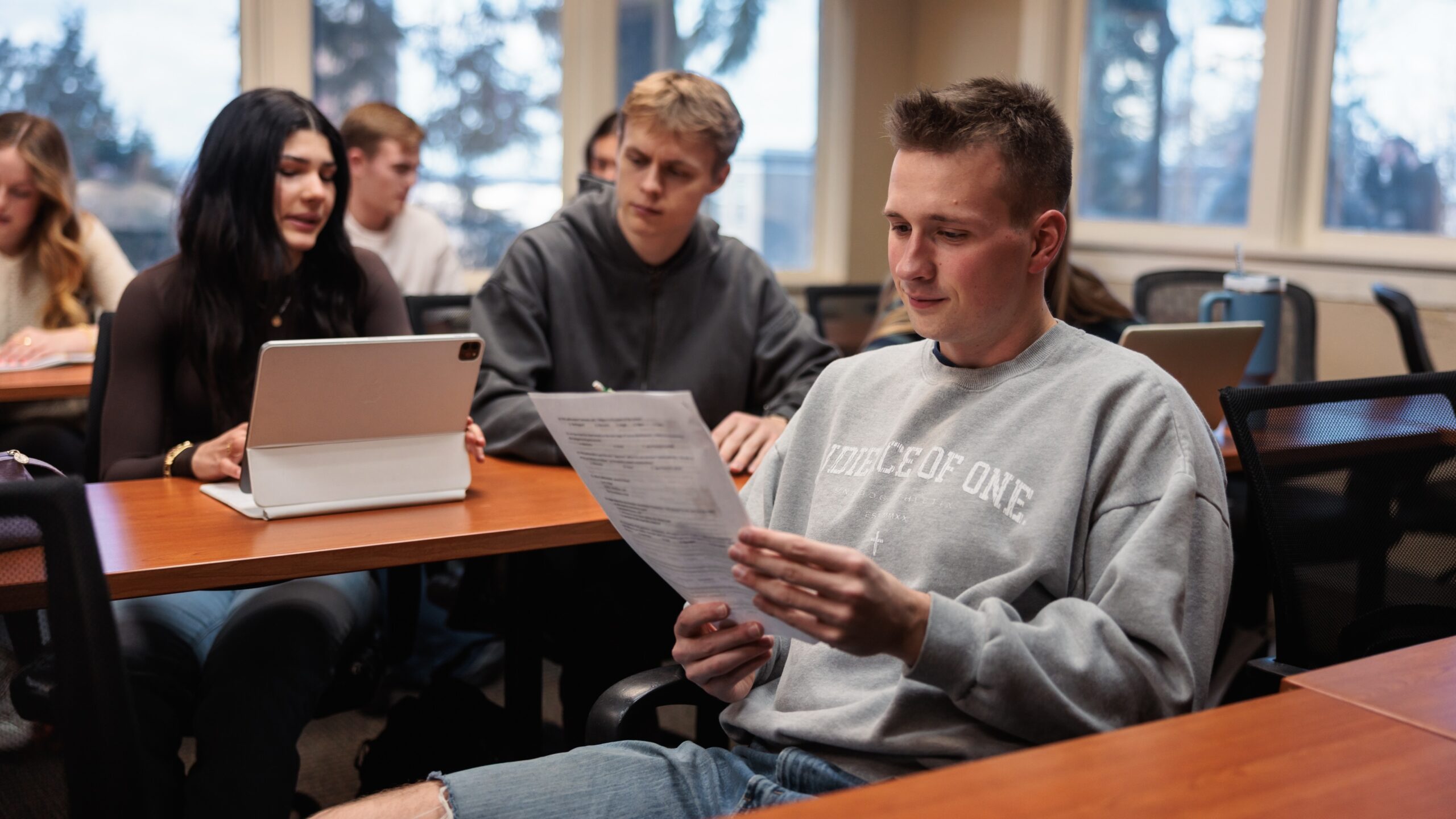 A group of students sit at tables in a classroom, focused on laptops and papers while engaged in a learning activity.