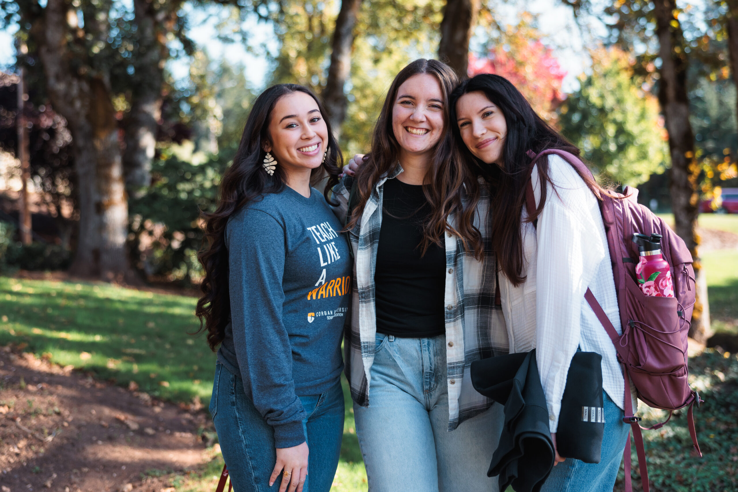 Three young women smiling and posing together surrounded by trees and greenery.