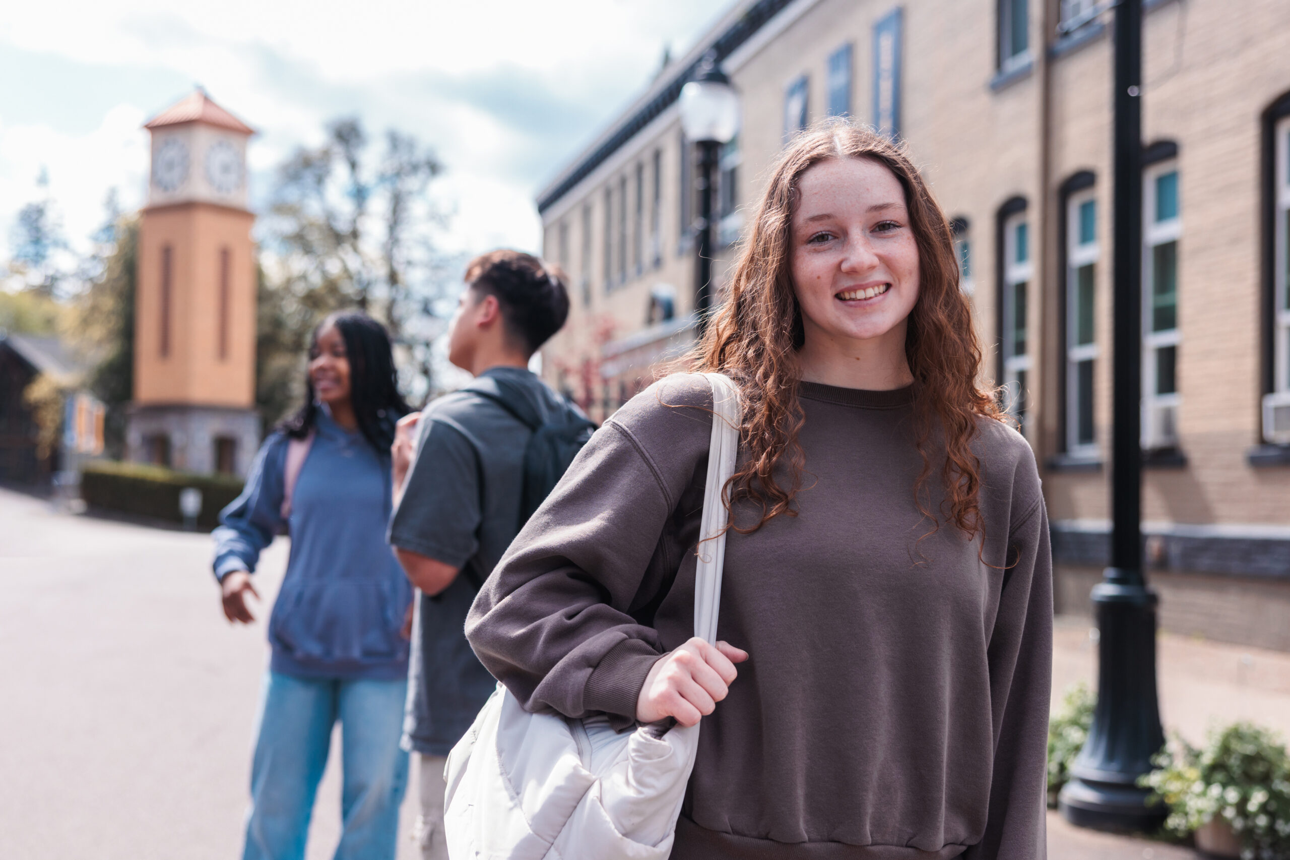 A young woman poses with a bookbag over her shoulder while her friends chat in the background on a college campus.