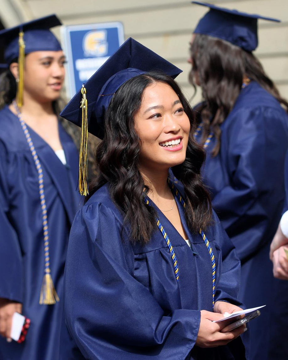 A young woman in a graduation cap and gown waits in line with a smile at Commencement