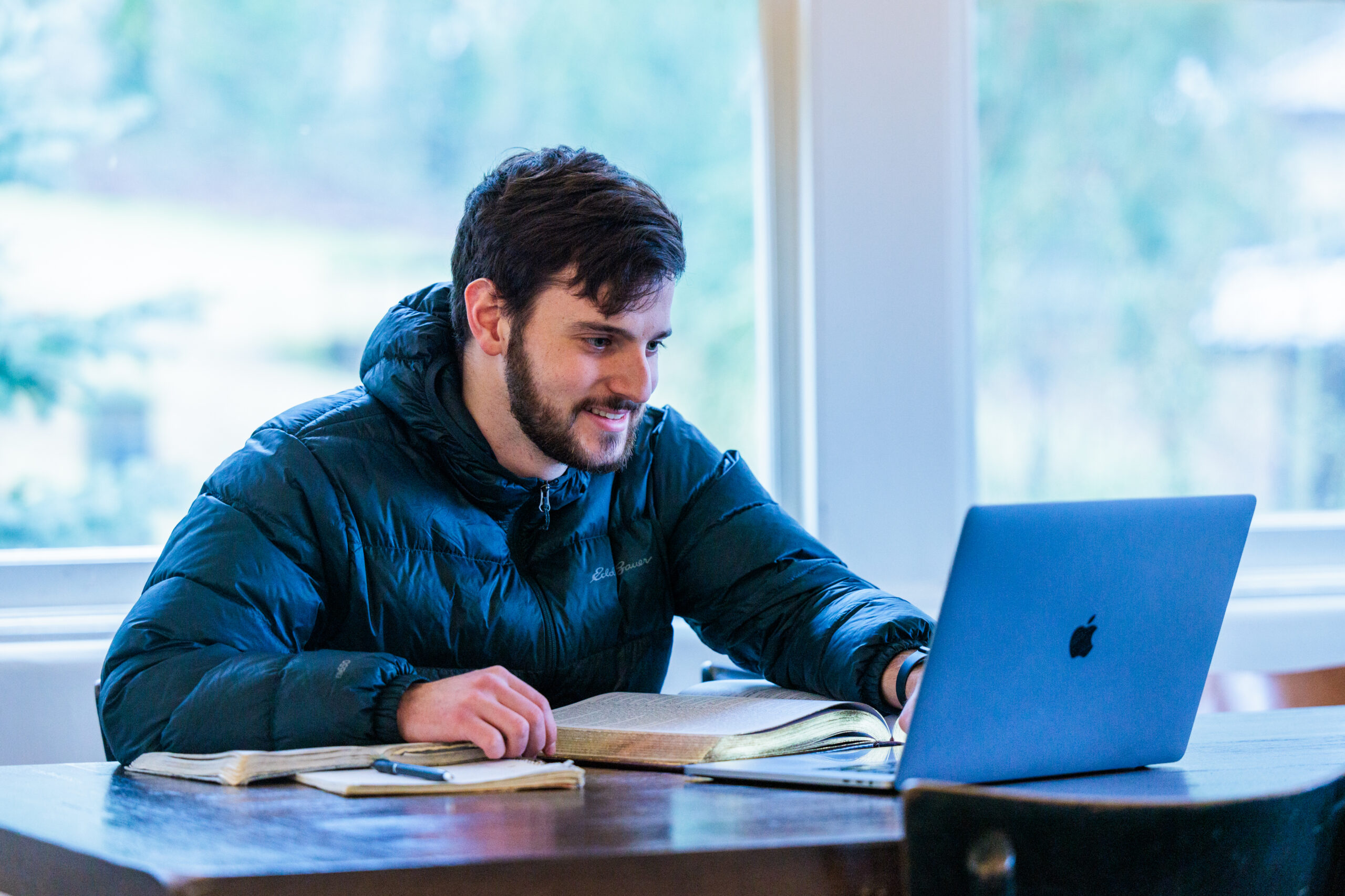 A man sits at a table engaged in an online class on his laptop with big, bright windows behind him.