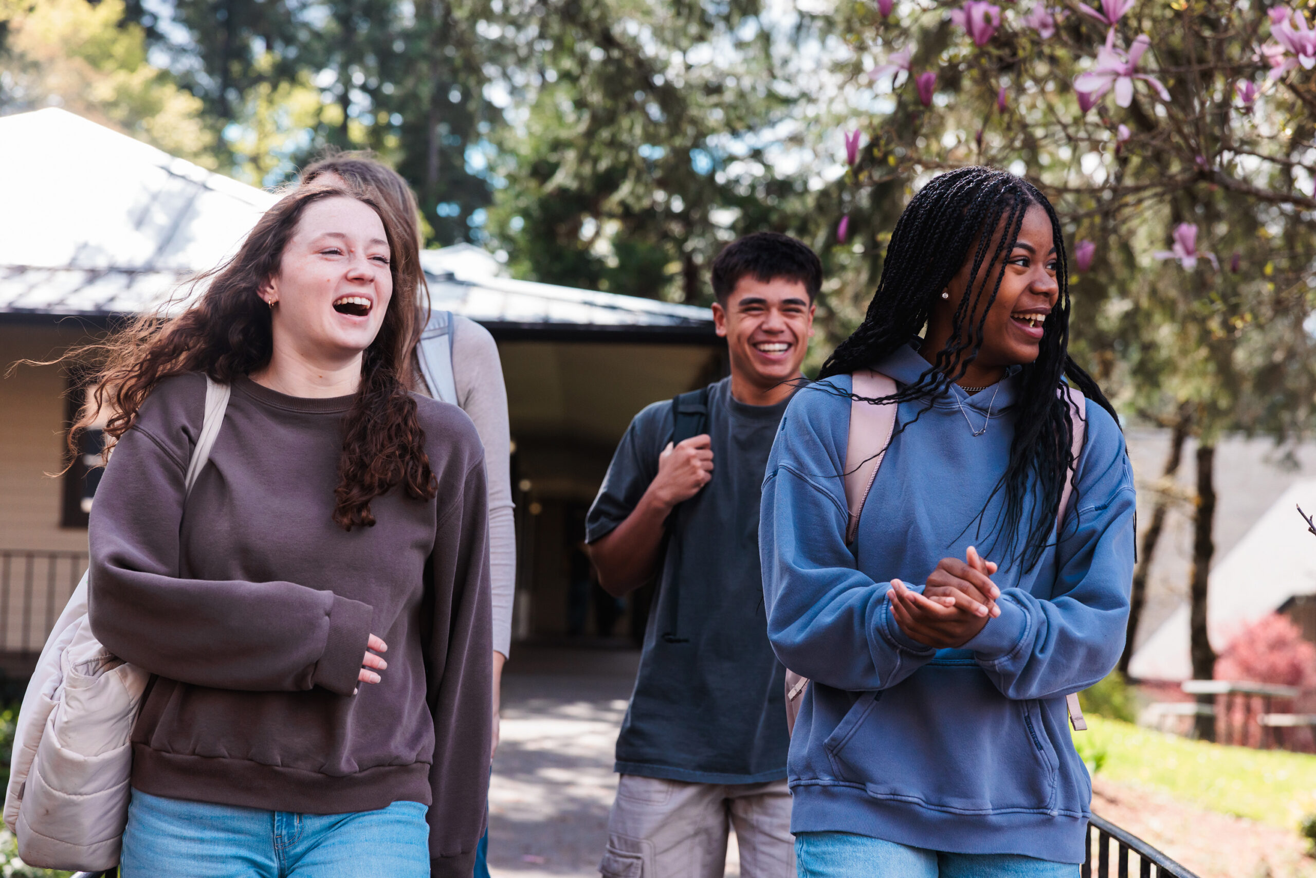 A group of four young friends walks side by side on a sidewalk, chatting and laughing on a bright day.