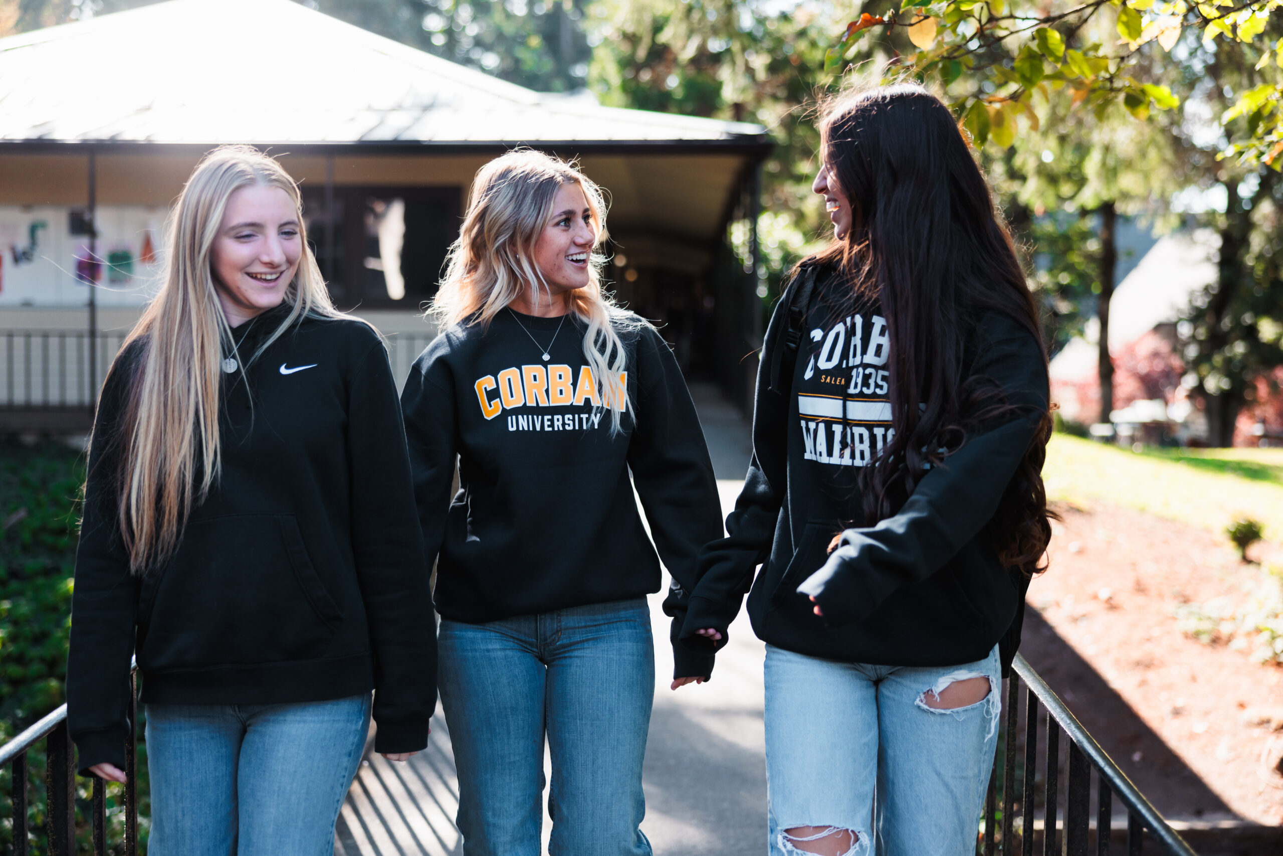 A trio of girls in black Corban sweatshirts and jeans walk side by side on a scenic path.
