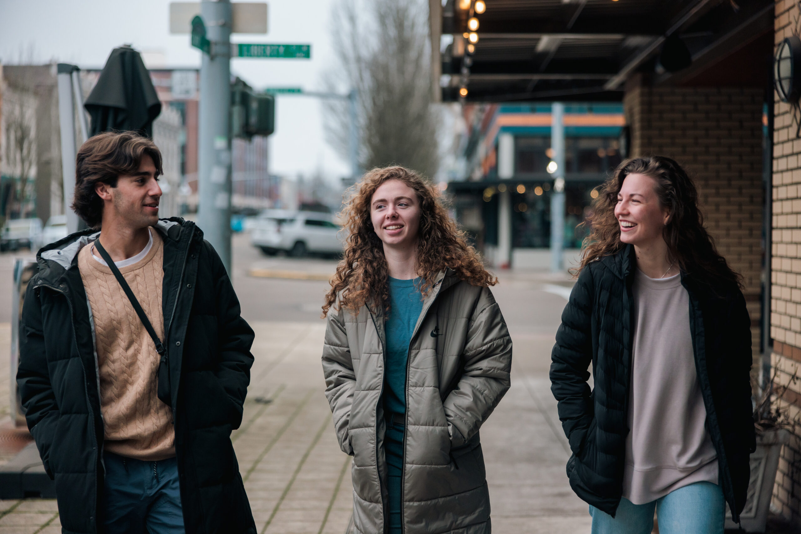 Three young people stand on a sidewalk in front of a building, smiling and chatting casually.