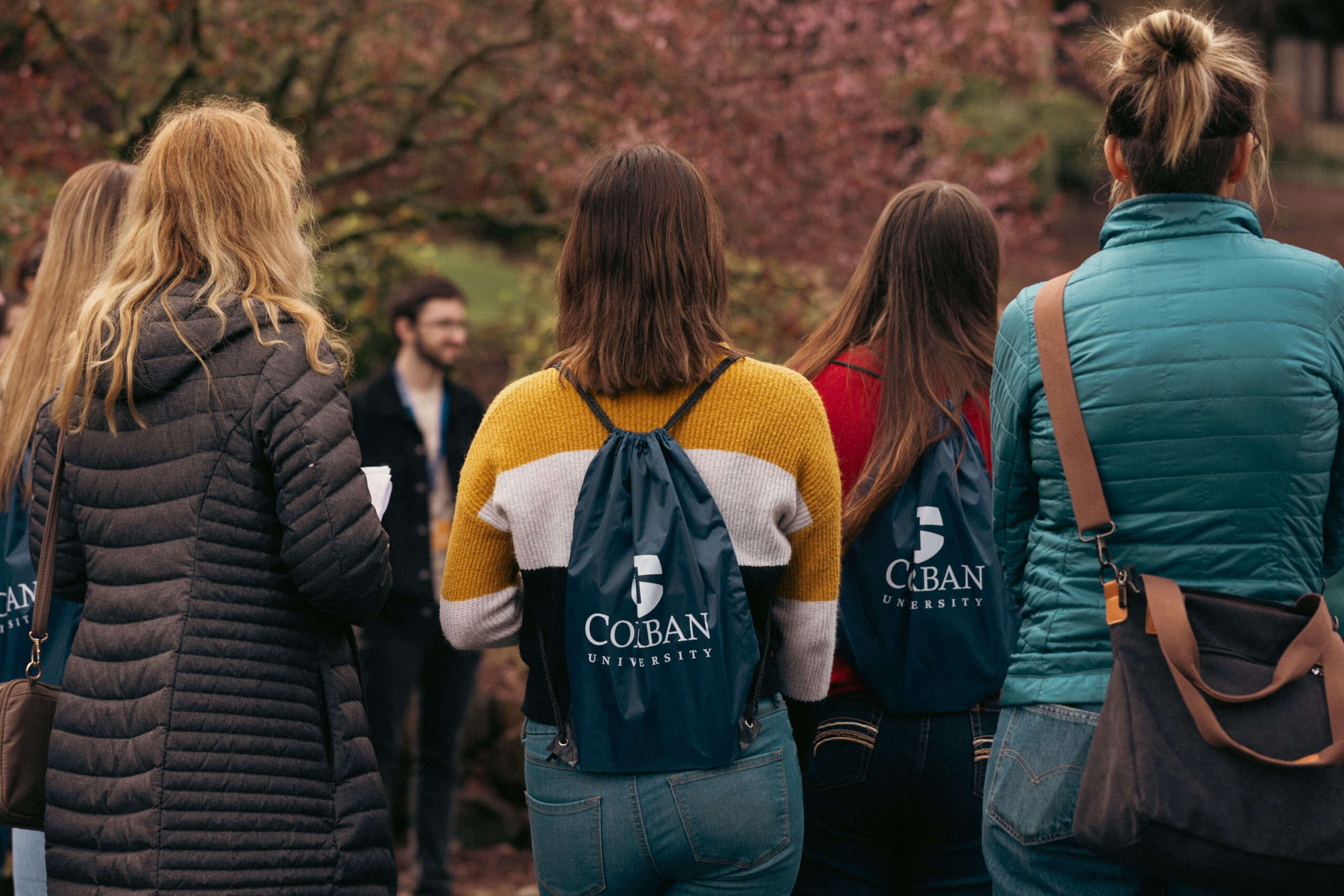 A group of people with backpacks stand outdoors facing away. Most wear jackets, and the backpacks have Corban University printed on them. The background is a blurred view of trees and a person facing the group.