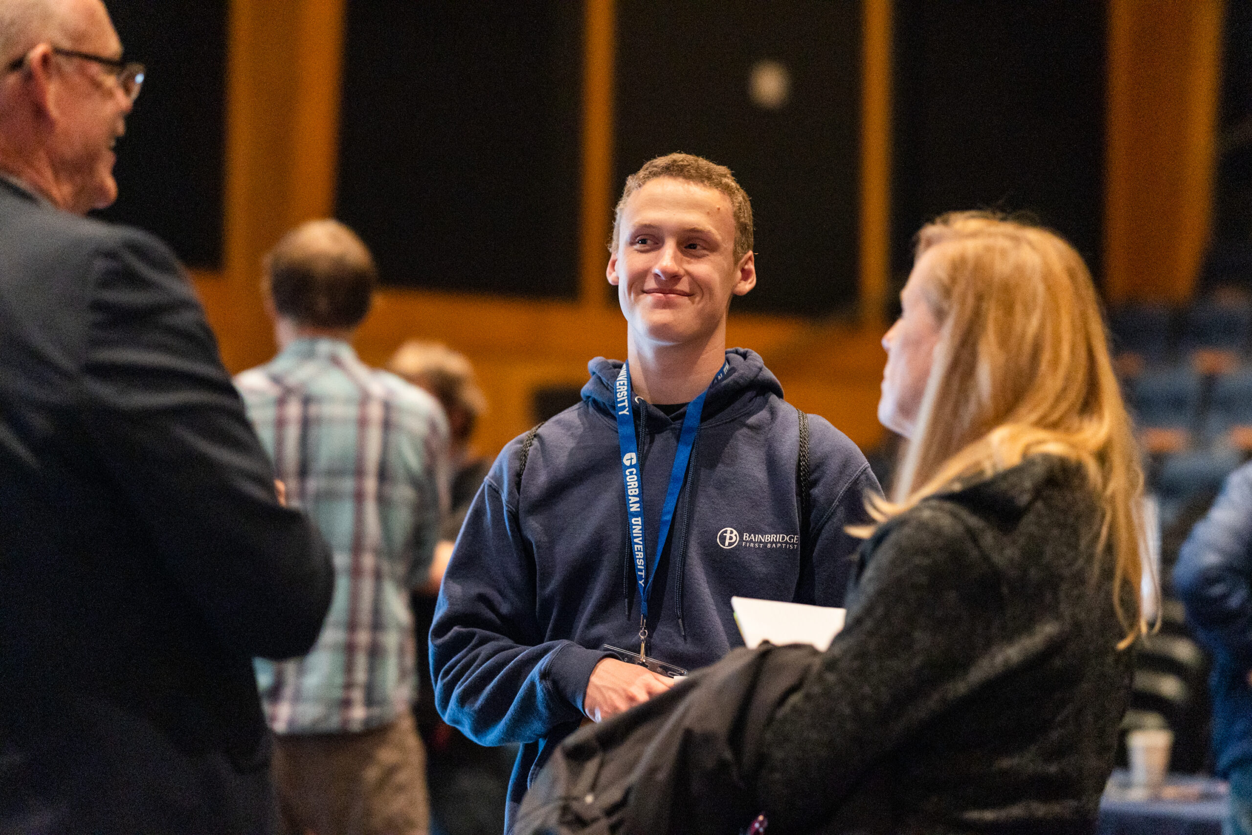 A young man wearing a navy hoodie and a lanyard smiles while engaging in conversation with two people. They are in a warmly lit indoor setting with blurred figures and seating in the background.
