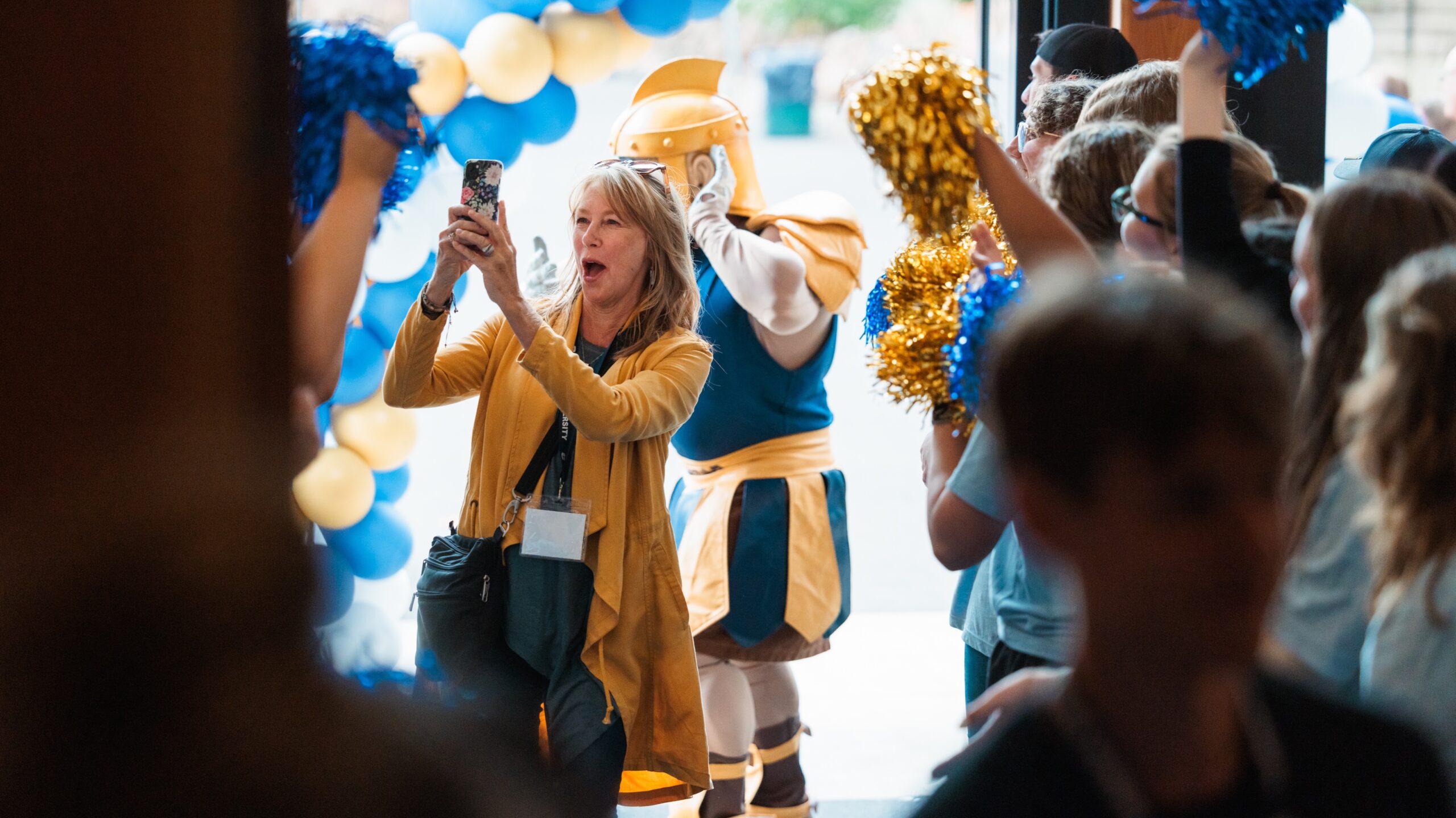 A woman cheers while recording on her phone as she walks through a group of cheering students.