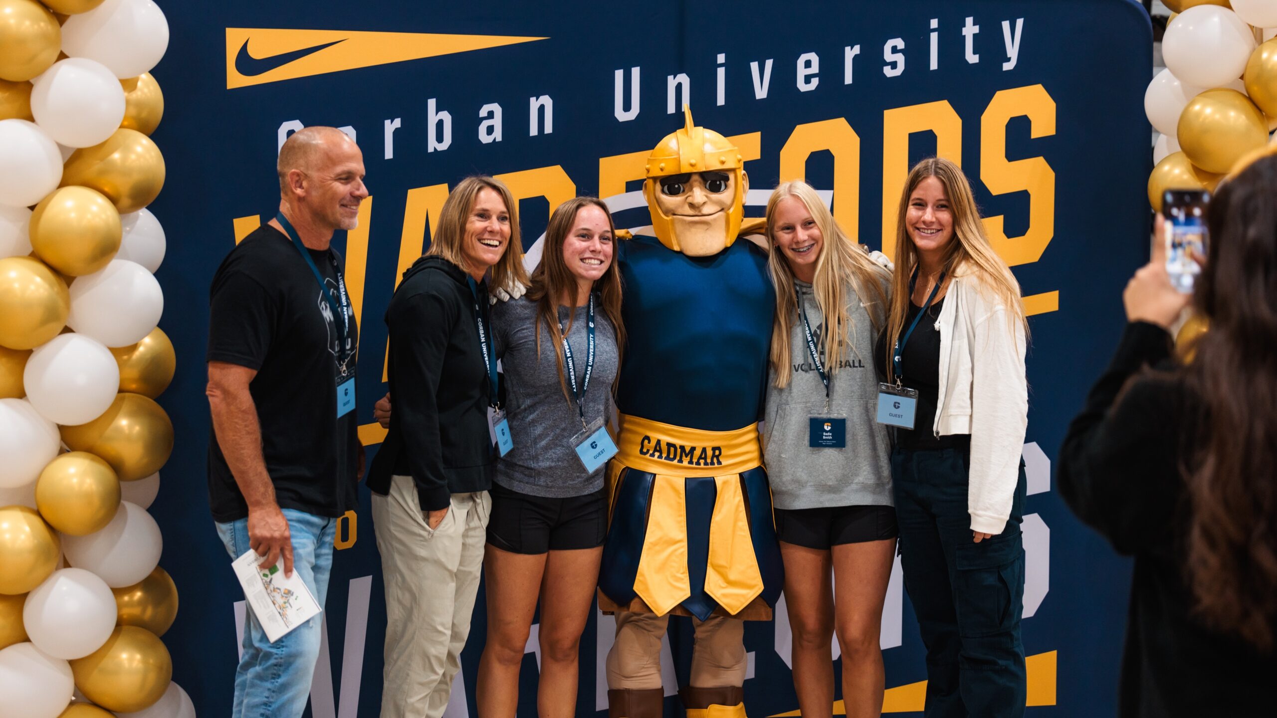 A group of five smiling individuals, including a person in a blue and yellow mascot costume, stand in front of a backdrop that reads Warriors. They are surrounded by white and gold balloons, and a person on the right is taking a photo of them.