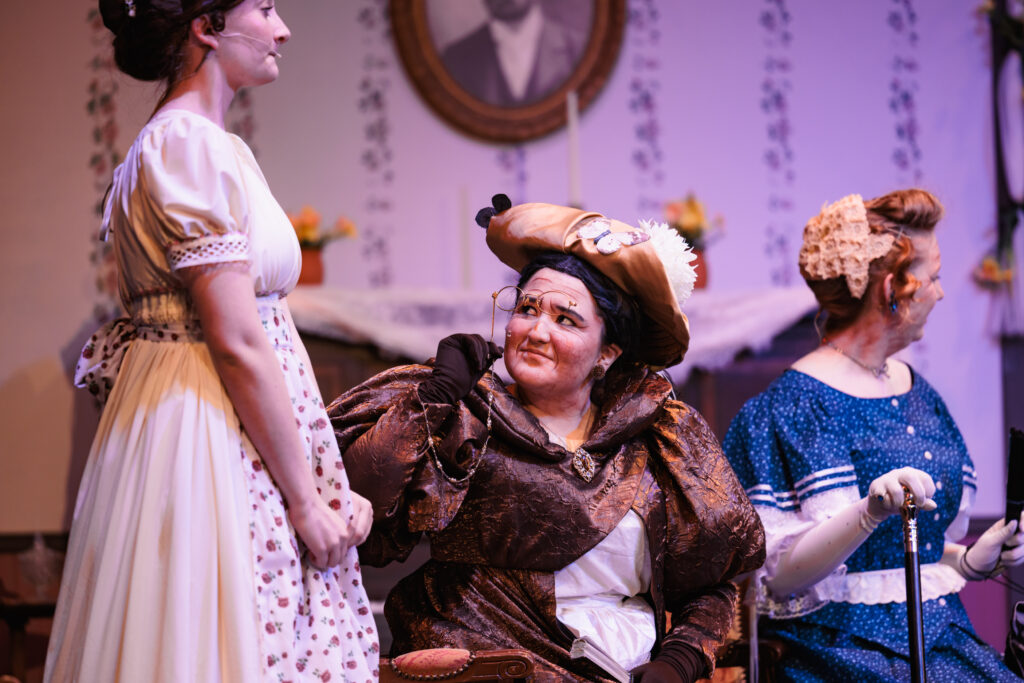 Three women in period dress seated on a stage, performing in the play "Good Wives."