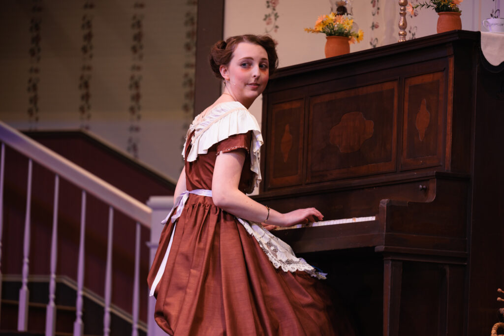 A woman in a dress playing a piano during a scene from the "Good Wives" play.