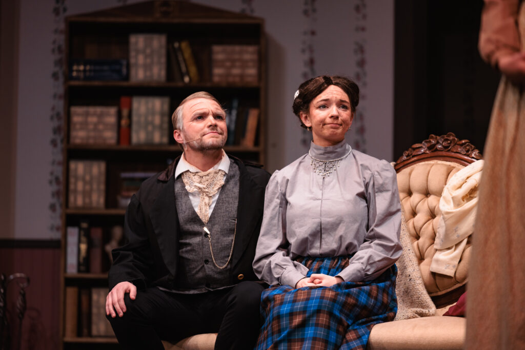 A couple sit on a couch in front of a bookcase, looking intently at their fellow actor for the "Good Wives" play.