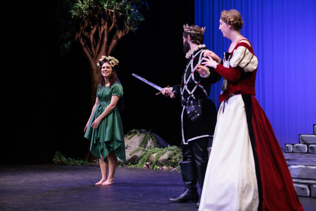 Three actors in costume perform on stage, featuring a tree backdrop, during the "Sleeping Beauty, Briar Rose" play.