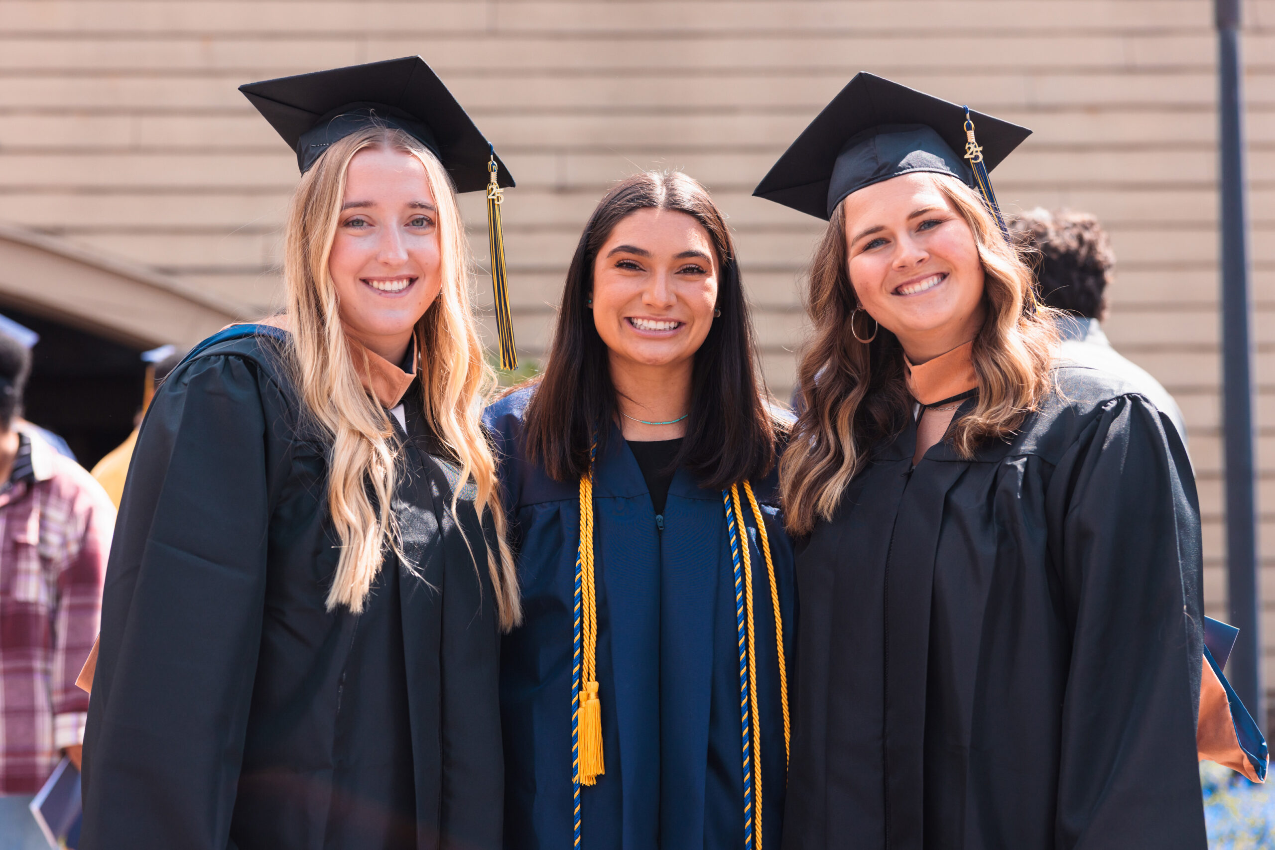 Three female graduates in caps and gowns smile together for a photo at Commencement 2025, celebrating their achievement