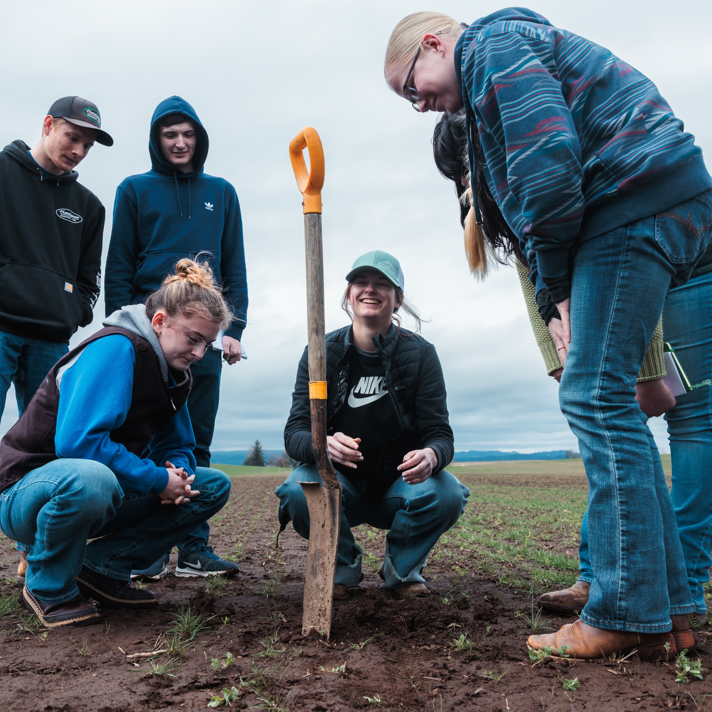 A group of students standing and kneeling in dirt around a shovel lodged upright in the soil with joyful expressions.