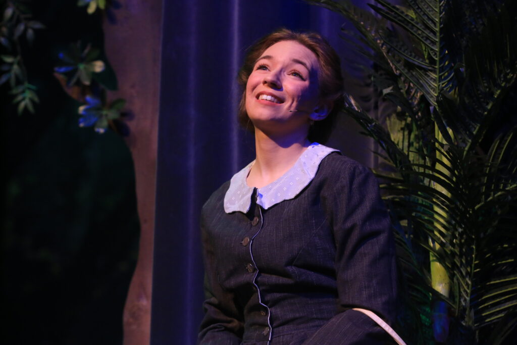 A woman in a stylish dress is sitting down, depicting a moment from the play "Around the World in 80 Days."