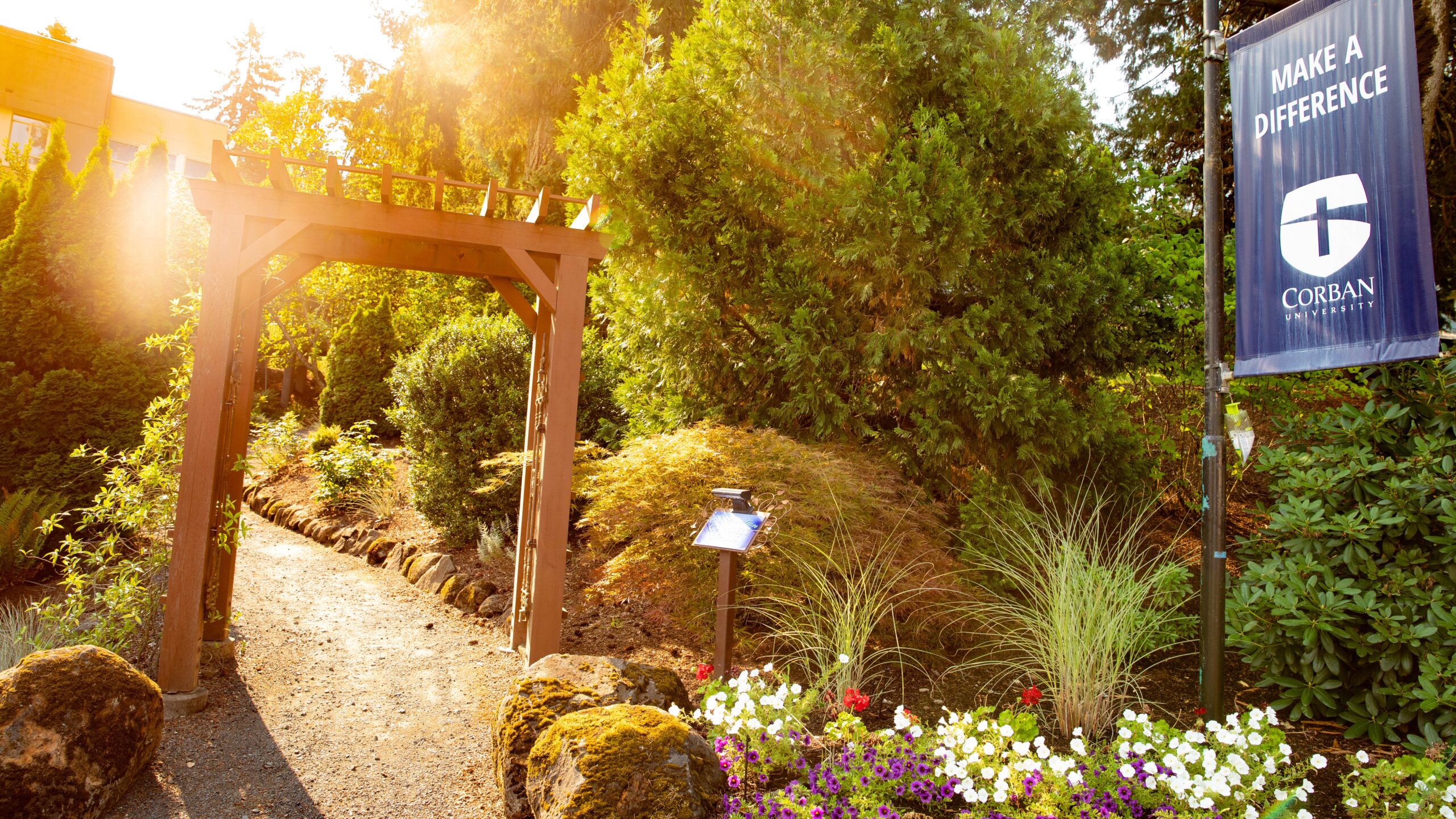 Sunlit garden path on Corban's Inspiration Garden Walk with a wooden archway surrounded by lush greenery and vibrant flowers.