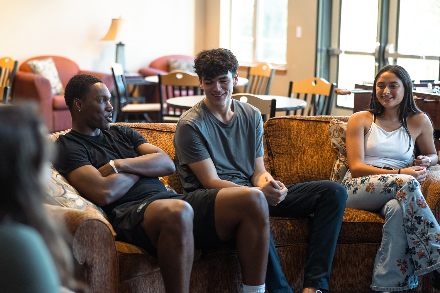 A group of friends sits together on a couch in a bright residence hall lobby, chatting and laughing.