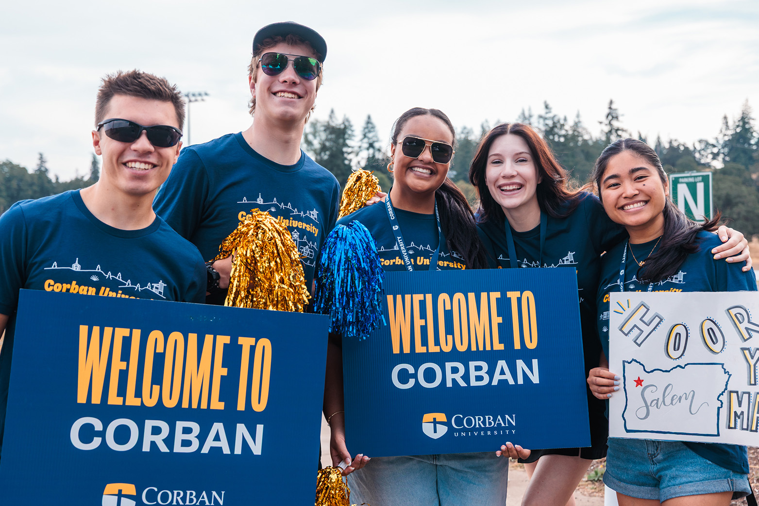 Four cheerful students stand by a welcome sign, ready for the event.