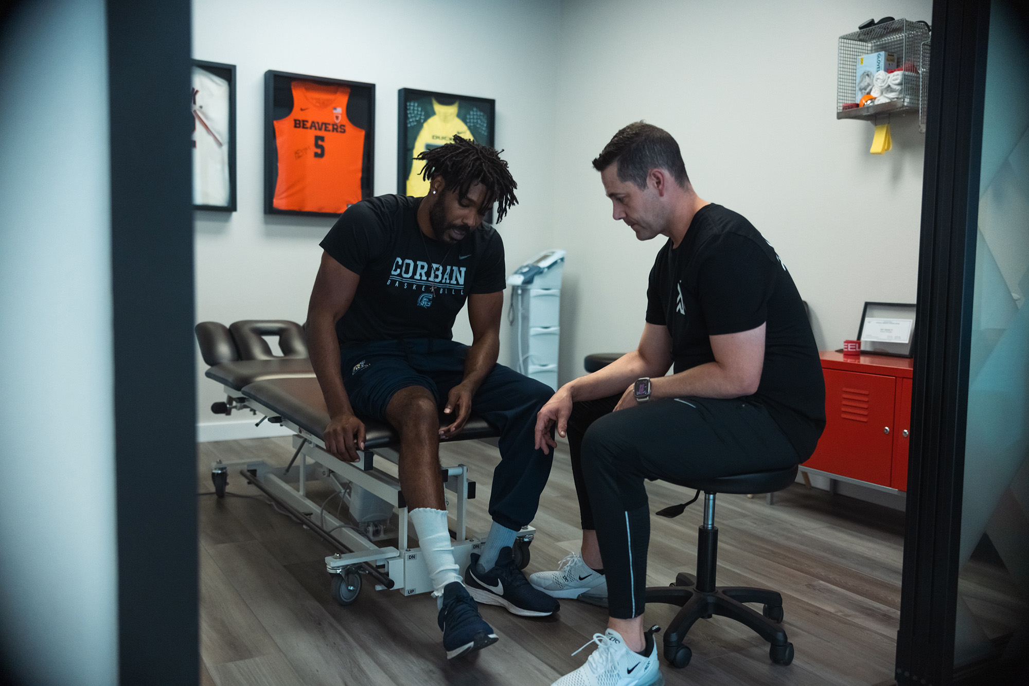 In a physical therapy room, a man sits on a bench while another man stands beside him, discussing treatment.
