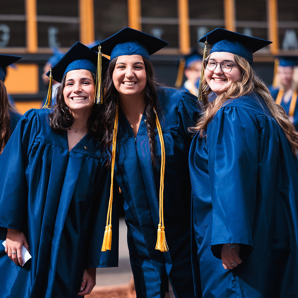 Three female graduates in dark blue caps and gowns smile on graduation day