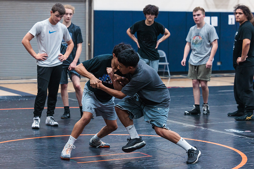 Two boys wrestle during a sports camp while other attendees look on.
