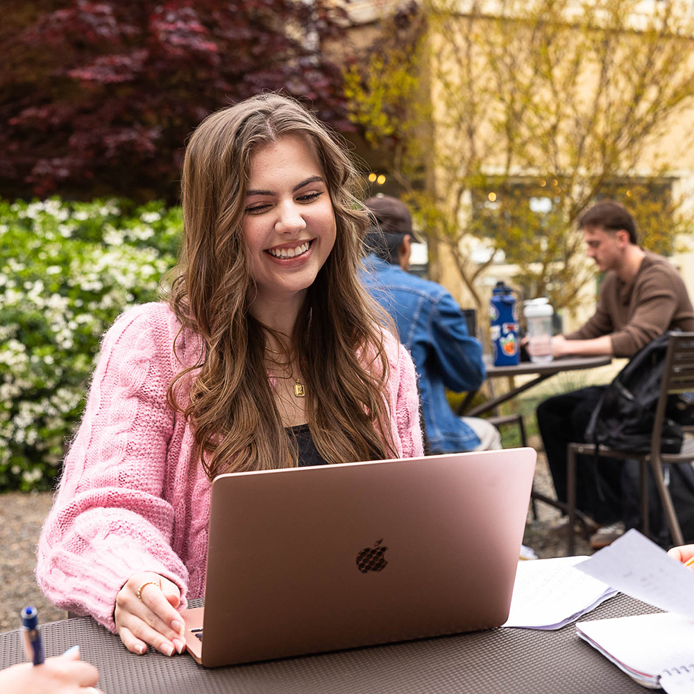 A female college student smiles while working on a laptop in an outdoor setting