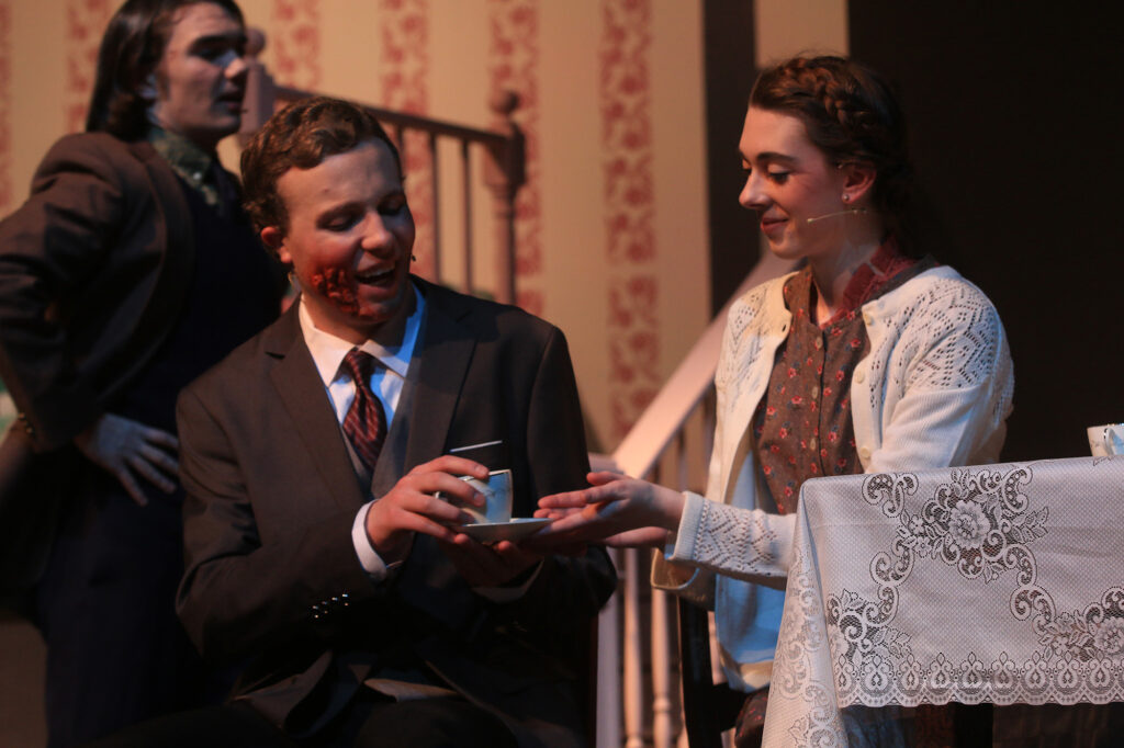 A woman passes a cup of tea to a man seated at a table in a home in a scene from The Hiding Place play