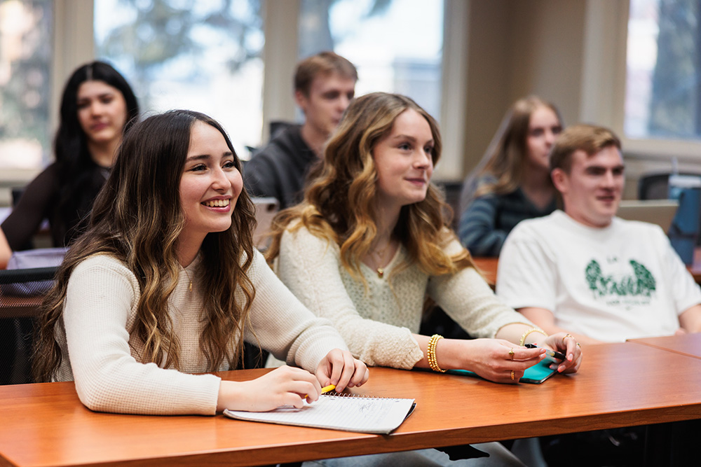 Several college students sit at tables, engaged in listening to a lecture in a classroom