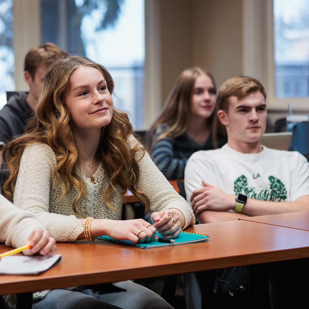 Several college students sit at tables, engaged in listening to a lecture in a classroom