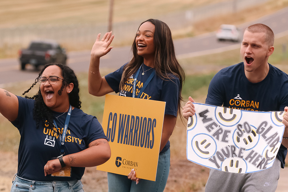 Three student leaders hold welcome signs and cheer for new students as they arrive at Warrior Welcome